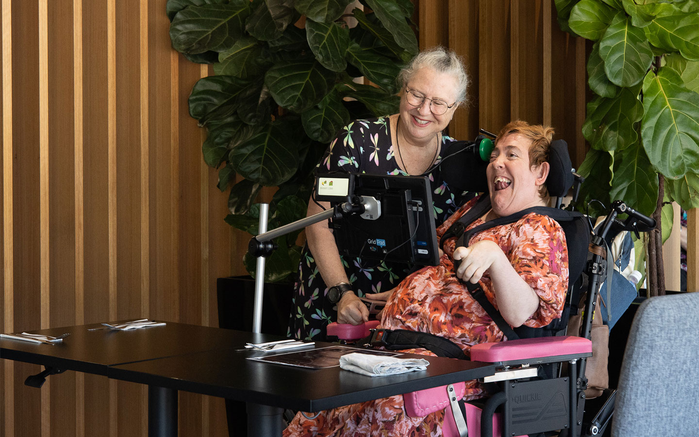 A smiling woman in a floral dress stands beside a woman in a wheelchair with a mounted communication device at a restaurant table. The atmosphere is warm and inviting, with wooden panel walls and large green plants in the background.